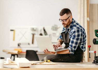 Gözlüklü ve önlüklü yetişkin erkek marangoz modern ışık işlerinde çalışırken laptopta bilgi arıyor.