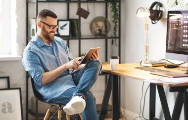 Günlük giysiler ve gözlük takan neşeli genç bir erkek iş yerinde büyük bir Monito ile kahve içerken tablet kullanıyor.