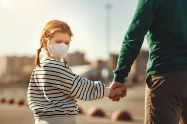 Back view of little girl in medical mask looking at camera over shoulder and holding hand of crop father while walking on parking lot during coronavirus outbrea clipart