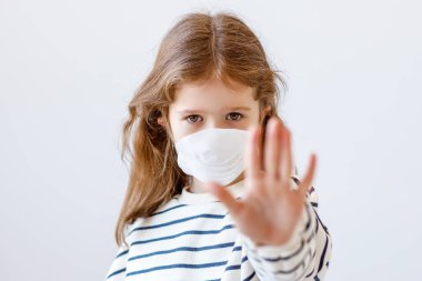 Tıbbi maskeli küçük bir kız kameraya bakıyor ve Coronavirus salgını sırasında gri arka grubuna karşı jest yapıyor.
