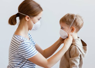 Coronavirüs salgını sırasında küçük bir çocuğa tıbbi maske takan yetişkin bir kadının gri arka grubuna karşı yan bakışı.