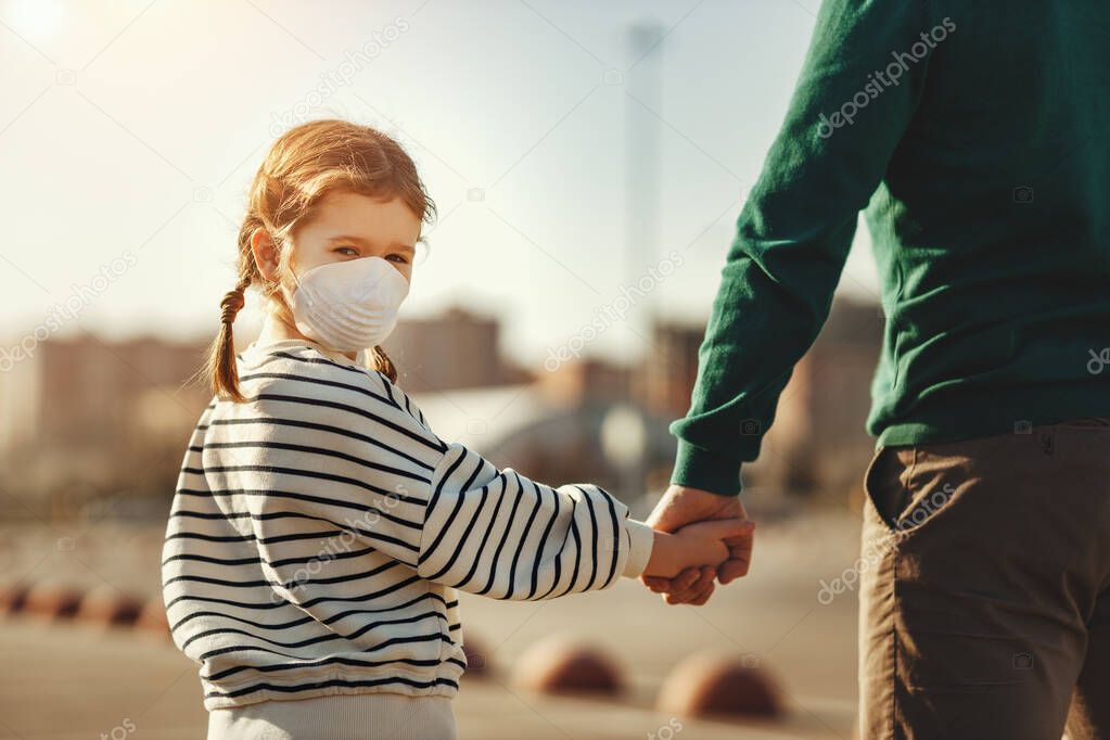 Back view of little girl in medical mask looking at camera over shoulder and holding hand of crop father while walking on parking lot during coronavirus outbrea
