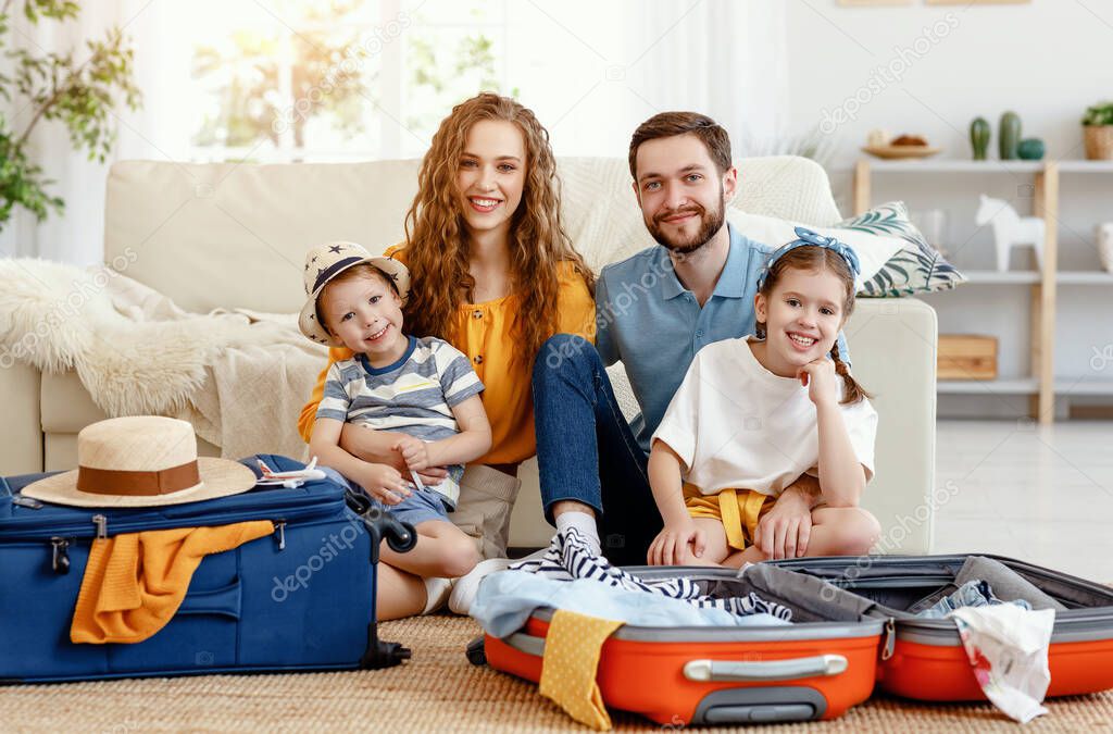 Happy parents with children laughing at camera while sitting on floor near sofa with suitcases near at hom