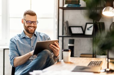 Günlük giysiler ve gözlük takan neşeli genç bir erkek iş yerinde büyük bir Monito ile kahve içerken tablet kullanıyor.