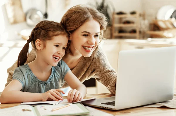 一位积极的年轻女性 一边帮助女儿在网上搜寻家庭作业的信息 一边与笔记本电脑一起坐在桌旁 — 图库照片