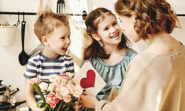 Cheerful little girl with card and youngest brother with  bouquet of tulip flowers smiling and congratulating happy mom on mother day at hom