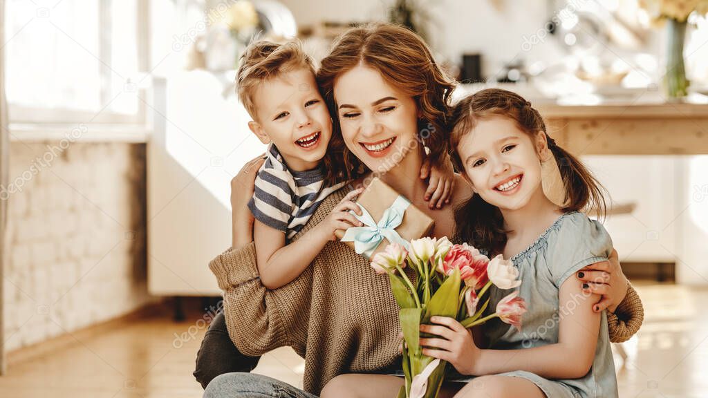 Cheerful little girl with bouquet of tulip flowers and youngest brother with gift box smiling and congratulating happy mom on mother day at hom