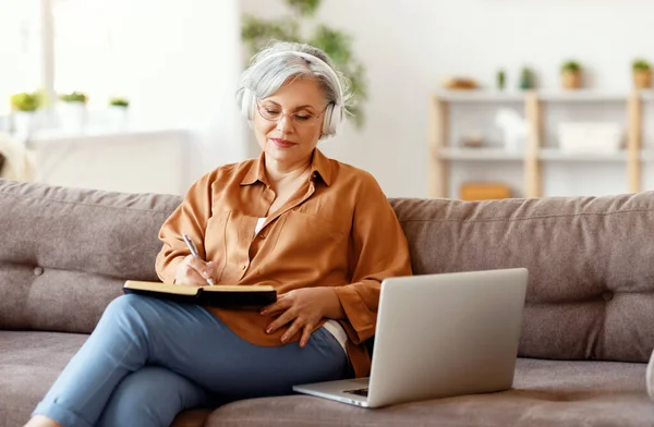 Oudere Vrouw Koptelefoon Bril Schrijven Notebook Het Luisteren Naar Muziek — Stockfoto