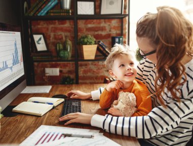 Gözlüklü genç anne, masada oturup bilgisayarında çizelgelerle çalışırken gülümseyen çocuğa bakıyor.