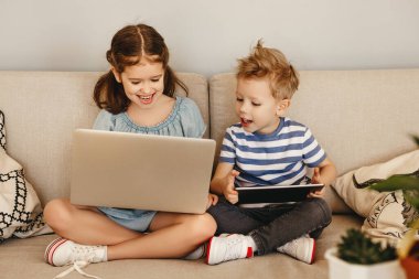 Mutlu küçük kız dizüstü bilgisayarda geziniyor ve en küçük kardeş evdeki kanepede birlikte otururken tablette oyun oynuyor.