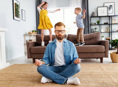 Rahat genç adam nilüfer pozisyonunda oturup meditasyon yaparken çocuklar evde kanepede zıplayıp eğleniyor.