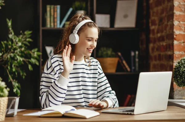 Joyful young female in wireless headphones waving wand greeting to screen while sitting at table and having video chat with business partners using laptop against blurred dark interior of comfortable loft offic