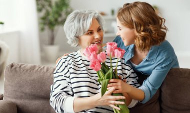Neşeli genç bayan, mutlu yaşlı anneye sarılıyor ve evde anneler gününü kutlarken bir buket lale veriyor.