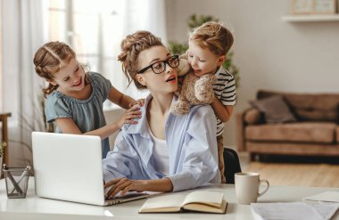 Kucaklaşan kız ve oyuncak gösteren küçük oğlan meşgul anne serbest çalışan masaya oturmuş bir fincan kahve içiyor ve dizüstü bilgisayarla aydınlık, modern dairelerin bulanık iç kısımlarına saldırıyor.