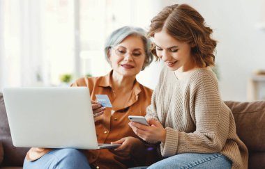 Kır saçlı genç bir kadın kanepeye dizüstü bilgisayarla oturuyor ve alışveriş sırasında kredi kartıyla online işlem yaparken akıllı telefon kullanıyor.