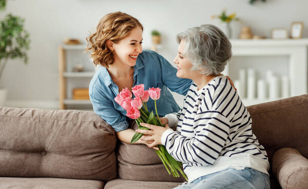Веселая молодая женщина обнимает счастливую пожилую мать и дарит букет тюльпанов, поздравляя с Днем матери в доме
