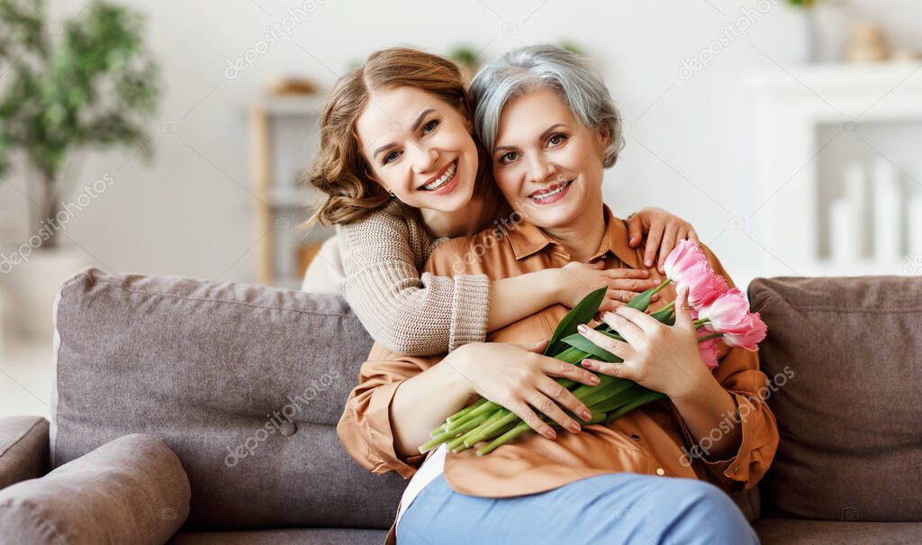 Cheerful young female smiling and embracing happy senior mother with bouquet of tulips in hands while sitting together on sofa in modern apartmen