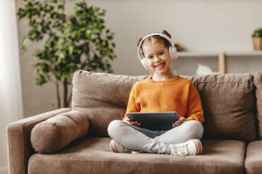 Evdeki yumuşak kanepede otururken günlük giysiler ve kulaklıklar takmış gülümseyen ve tablette oyun oynayan neşeli bir kız.