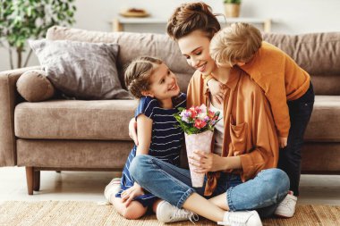 Neşeli, rahat anne kanepenin yanında oturmuş renkli taze çiçekler sunarken küçük kız ve oğul anneler günü tatilinde kucaklaşıp tebrik ediyorlar.