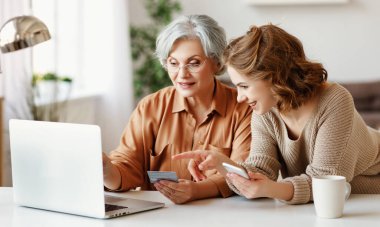 Akıllı telefonlu genç bir kız, evde internetten alışveriş yaparken bilgisayarında yaşlı bir bayana gülümsüyor ve bilgisayarını gösteriyor.
