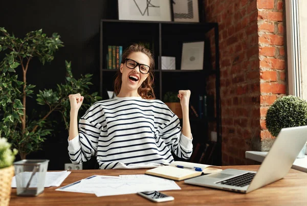 Happy young lady with eyes closed and arms raised rejoicing in success while sitting at wooden table with notepad and laptop in loft office among green potted houseplant