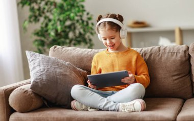 Evdeki yumuşak kanepede otururken günlük giysiler ve kulaklıklar takmış gülümseyen ve tablette oyun oynayan neşeli bir kız.