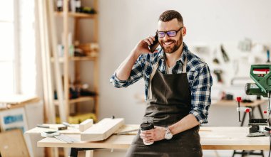 Önlüklü, elinde kahve bardağıyla mutlu genç bir adam akıllı telefondan konuşuyor ve modern atölyede tezgaha yaslanıyor.