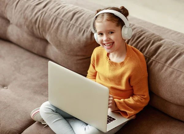 从头上高高兴兴的小女孩手中接过耳机 微笑着看着摄像机 双腿交叉坐在沙发上 在客厅里用笔记本电脑 — 图库照片