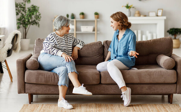 Full length young woman smiling and talking with senior mother while sitting on comfortable sofa at home togethe