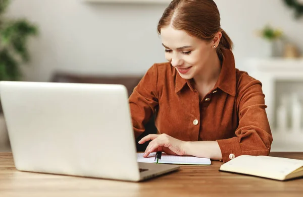 机に座って家でリモートプロジェクトに取り組んでいる間 ノートパソコンやノートパソコンを読んで茶色のシャツを着た陽気な女性 — ストック写真