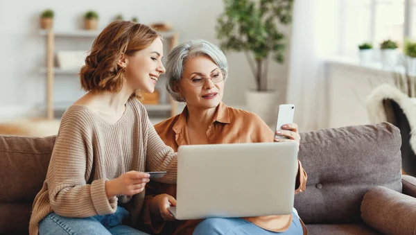 Kır saçlı genç bir kadın kanepeye dizüstü bilgisayarla oturuyor ve alışveriş sırasında kredi kartıyla online işlem yaparken akıllı telefon kullanıyor.