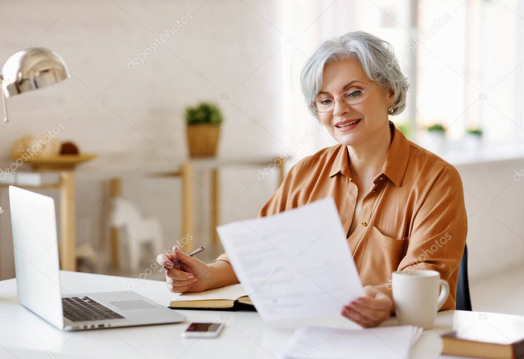 Senior woman with laptop and notebook  while sitting at table and working on remote project at hom