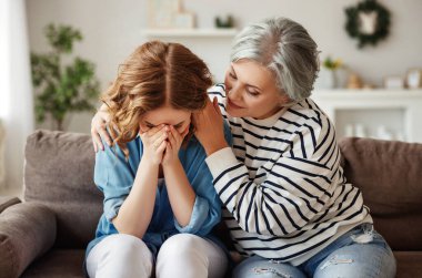 Yaşlı bir kadın, evde kanepede birlikte otururken ağlayan küçük kızını kucaklıyor ve destekliyor.