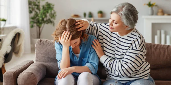 Elderly woman embracing and supporting crying young daughter while sitting on couch at home togethe