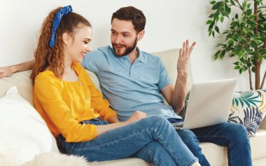 Sakallı adam, dizüstü bilgisayarlı, gülümseyen kız arkadaşıyla el kol hareketleri yapıp sohbet ederken evdeki rahat oturma odasında birlikte oturuyor.