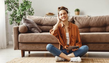 Gündelik kıyafetleri ve kablosuz kulaklıkları olan genç bir kadın. Dijital tablet müzik dinlerken, modern oturma odasındaki yumuşak kanepenin yanında, bacak bacak bacak üstüne atmış bir şekilde oturuyordu.