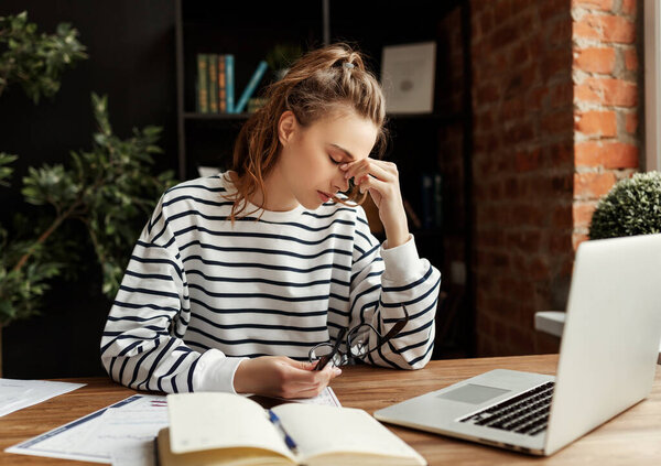 Frustrated student in casual clothes with eyes closed trying to solve problem while sitting at table with notepad and laptop and touching head in modern offic