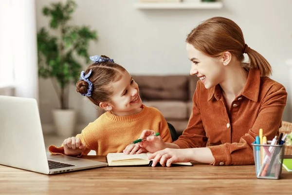 一位积极的年轻女性 一边帮助女儿在网上搜寻家庭作业的信息 一边与笔记本电脑一起坐在桌旁 — 图库照片