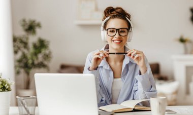 Gözlüklü ve kablosuz kulaklıklı kendini işine adamış bir kadın kameraya gülümserken elinde bir kupa sıcak içecekle dizüstü bilgisayarın başında ışık dolu modern yaşam kangurusuyla not defterine yazı yazıyor.