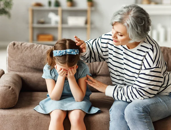 Elderly woman in casual clothes embracing and comforting crying girl while sitting on sofa at home togethe