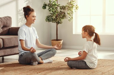 Beyaz tişörtlü ve gri taytlı mutlu kadın ve kız lotus poz verip birbirlerine bakıyorlar ve yog çalıyorlar.