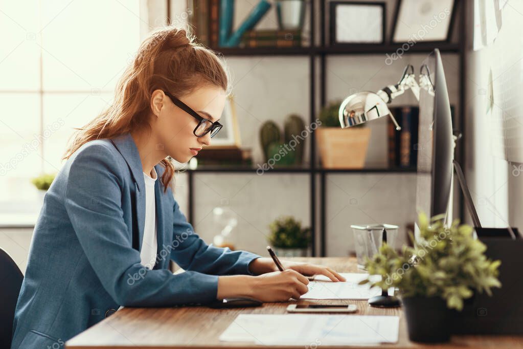 Side view of serious concentrated young female specialist sitting at desk and making notes in document while working at workplac