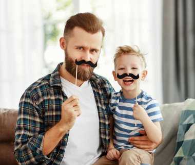 Mutlu genç sakallı adam ve küçük oğlu evdeki kanepede birlikte otururken eğleniyor ve takma bıyıkla oynuyorlar.