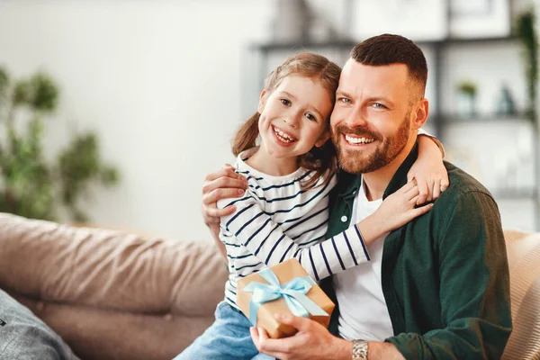 Happy young man with gift in hand and adorable little daughter embracing and looking at camera while sitting together on sofa in cozy living room on fathers da