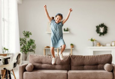 Uzun boylu, kulaklıklı, gülen ve kollarını kaldırarak kanepede zıplayan müzik dinleyen ve evde eğlenen bir kız.