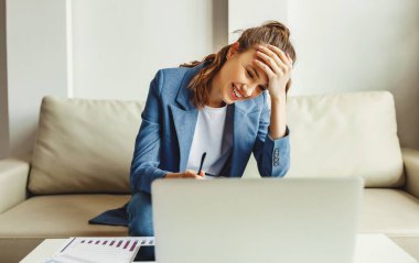 Genç bayan girişimci gündelik kıyafetleriyle koltukta oturmuş gülerken ve laptopta iyi haberler okurken.