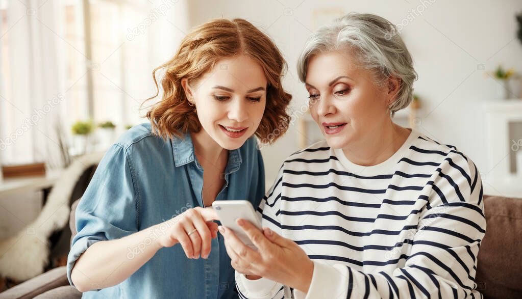 Happy senior woman smiling and sharing data on smartphone with young daughter while sitting on couch at home togethe