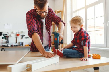 Günlük giysiler içinde neşeli küçük bir çocuk gülerek ve evde atölyede mobilya montajı yaparken babasının delik açmasına yardım ederek