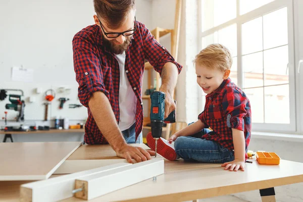 Delighted little boy in casual clothes laughing and helping father to drill hole while assembling furniture on table in home workshop togethe