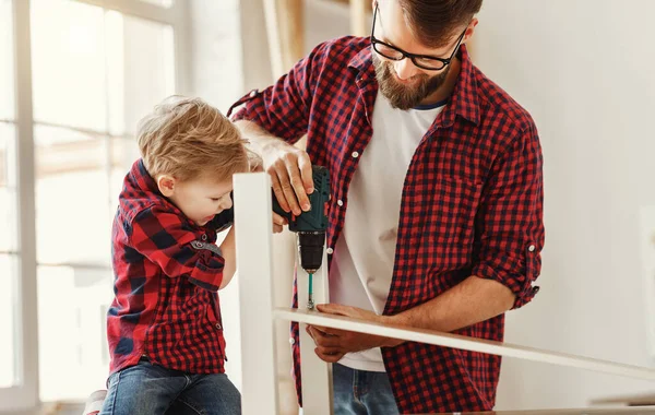 Delighted little boy in casual clothes laughing and helping father to drill hole while assembling furniture on table in home workshop togethe